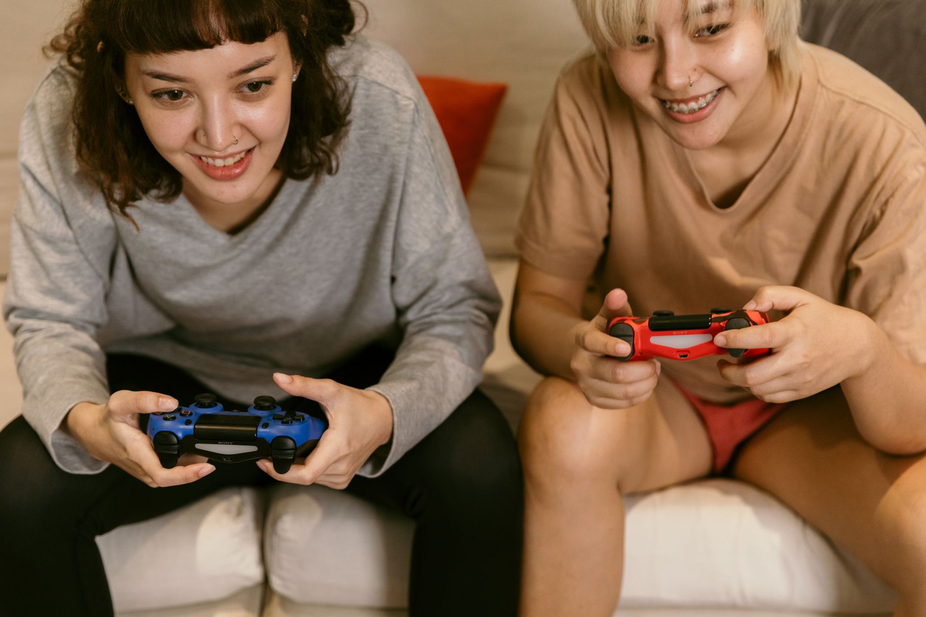 Low angle of cheerful crop Asian females in home clothes playing video games with joysticks in hands on sofa in living room —